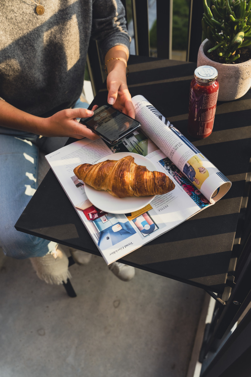 Eine Person sitzt an einem kleinen Tisch auf dem ein Croissant und ein Magazin liegt mit einem Smartphone in der Hand 