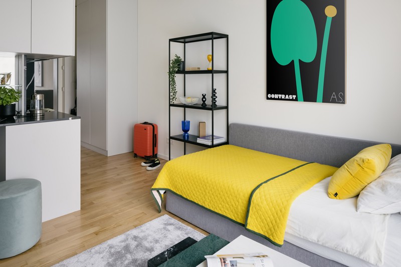 Mikroapartment in Berlin und Bett mit gelber Tagesdecke, schwarz-türkisem Bild und Regal an der Wand