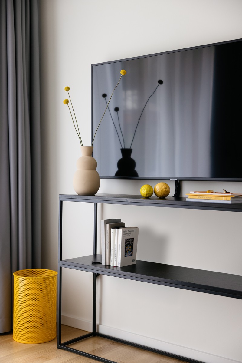 Black shelf with vase, books and magazines on it. Next to it a yellow wastebasket, above it a TV on the wall 