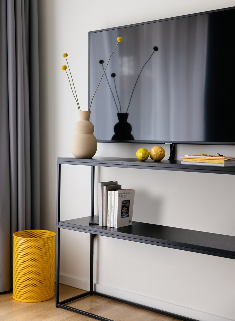 Black shelf with vase, books and magazines on it in our Business Apartments in Berlin. Next to it a yellow wastebasket, above a TV on the wall 