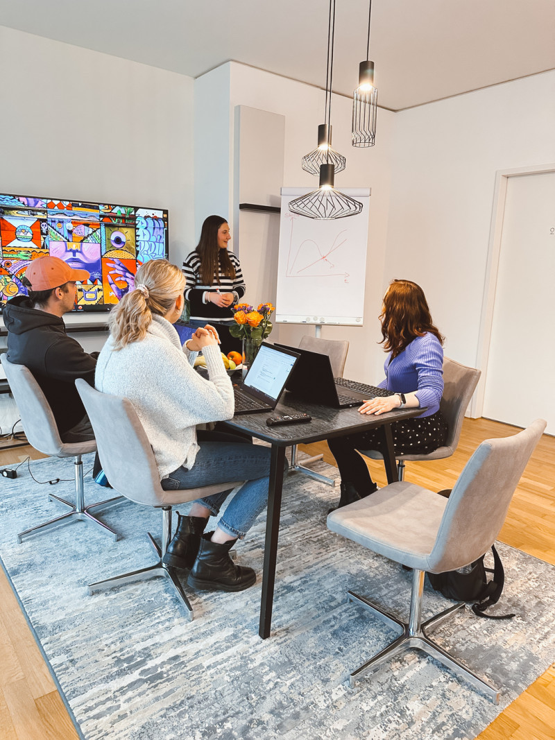  Ein Büro mit vier Personen, die an Laptops arbeiten und ein TV-Bildschirm im Hintergrund.