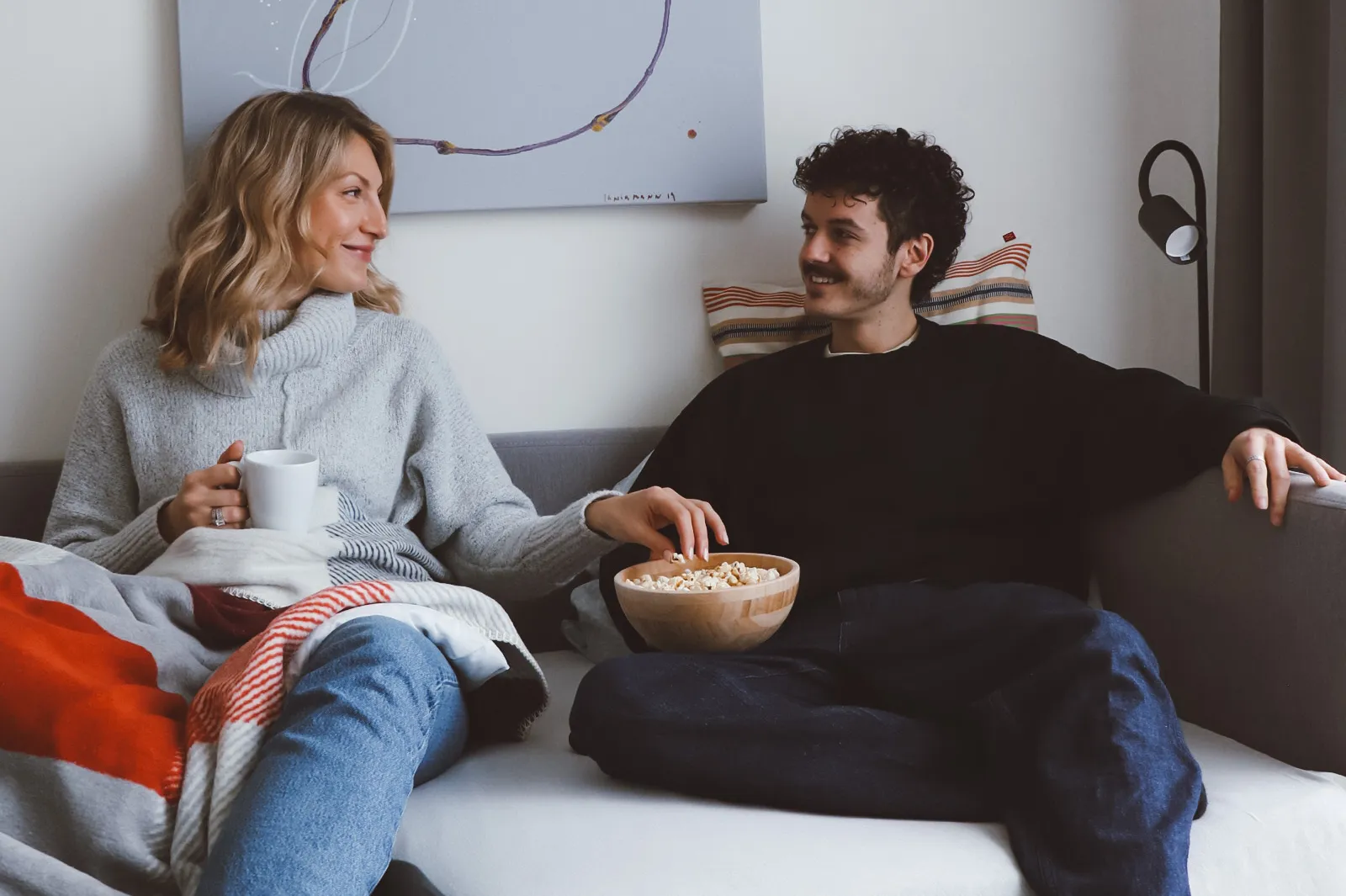 Ein Paar sitzt gemütlich auf dem Sofa, in eine Decke gehüllt, und teilt sich eine Schüssel Popcorn. Sie lächeln sich liebevoll an – ein intimer, entspannter Moment für einen ruhigen Abend zu Hause.