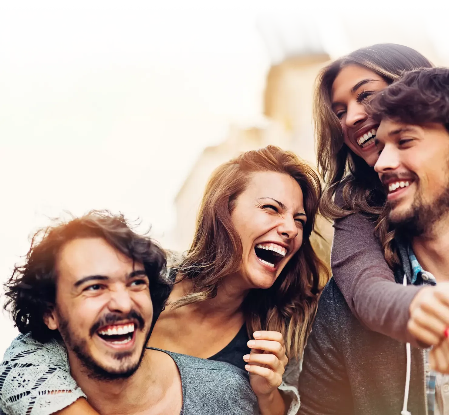 Four young friends are laughing joyfully outdoors, clearly sharing a fun and happy moment together. They are close, smiling broadly, and appear carefree. The warm tones and blurred background suggest a sunny day in a city or town.