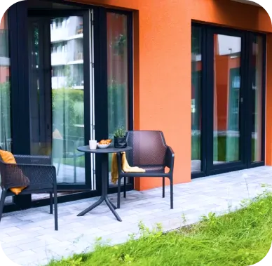 Small private terrace in front of an apartment with a vibrant orange exterior. Two black chairs and a round table are set on a stone patio, creating a cozy outdoor seating area next to a grassy lawn.