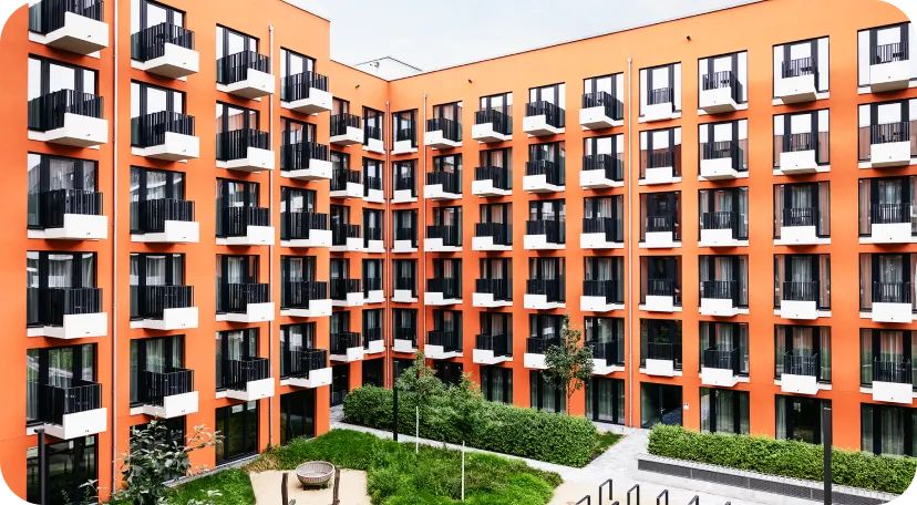 Modern residential building with a bright orange facade, featuring multiple identical balconies and large windows. The inner courtyard below includes green landscaping with trees, bushes, and a circular seating area.
