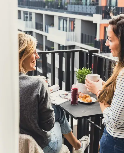 Two women enjoying a relaxed conversation on a modern balcony, sipping coffee and sharing a breakfast with croissants, against the backdrop of a contemporary apartment complex.