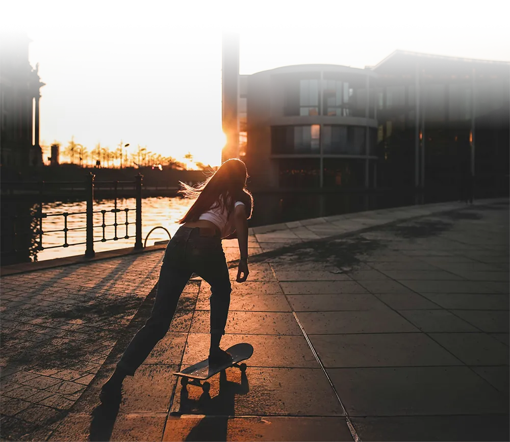 Silhouetted person pushing a skateboard along a riverside promenade at sunset, with modern buildings and warm sunlight in the background creating a vibrant, urban atmosphere.