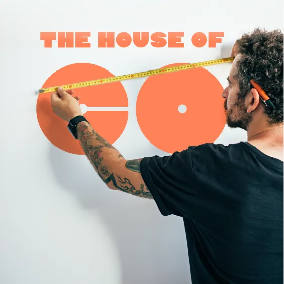 A man with tattoos on his arm and a pencil behind his ear is measuring orange geometric shapes on a white wall with a yellow tape measure. Above the shapes is the bold red text “THE HOUSE OF CO”. The image conveys creativity and precision in a co-living or design context.