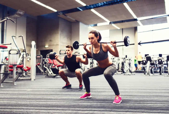 A woman and a man are performing squats with barbells on their shoulders in a modern gym. Both are wearing athletic clothing, and the gym is filled with fitness equipment and machines in the background. The scene emphasizes strength training and a healthy lifestyle.