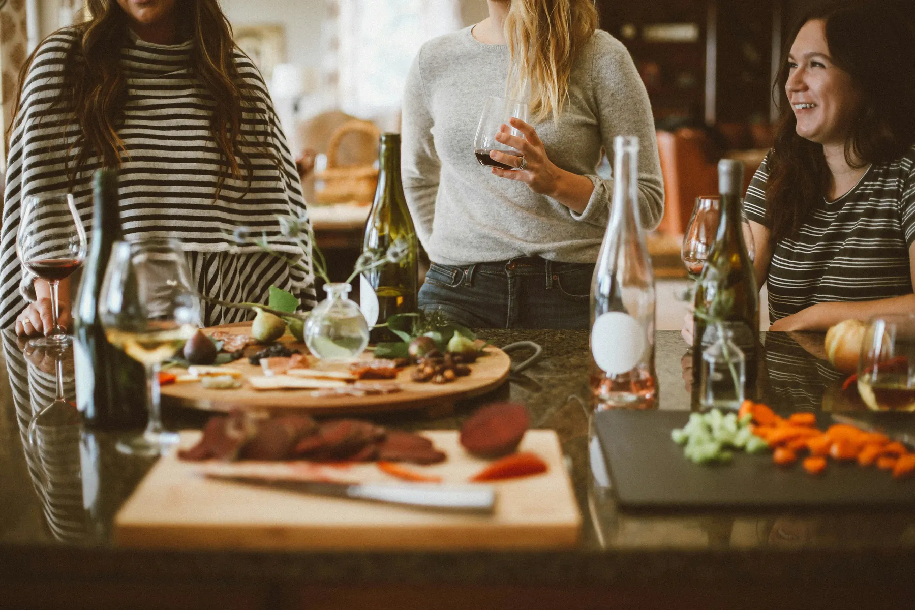 A group of women casually gathered in a cozy kitchen, enjoying wine and conversation. A rich assortment of food like sliced meat, figs, and vegetables is laid out on cutting boards and a counter, suggesting a relaxed social evening or dinner prep.