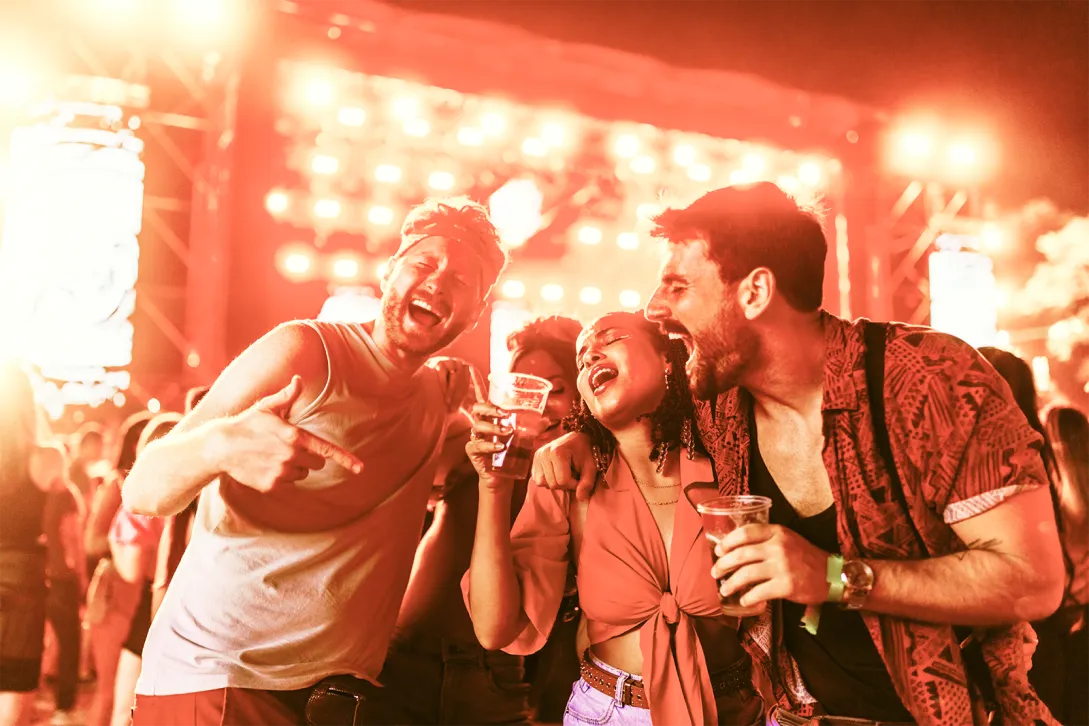 Three young adults enthusiastically celebrate and sing along at a vibrant music concert or festival. They're smiling, holding drinks, and enjoying the energetic atmosphere illuminated by bright, colorful stage lights. Their expressions and body language convey excitement, joy, and carefree fun.