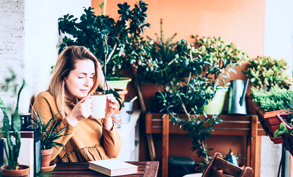 A woman sits peacefully at an outdoor wooden table surrounded by numerous green plants. She enjoys a quiet moment sipping from a white mug with eyes closed, a book on the table in front of her, creating a serene and tranquil mood.