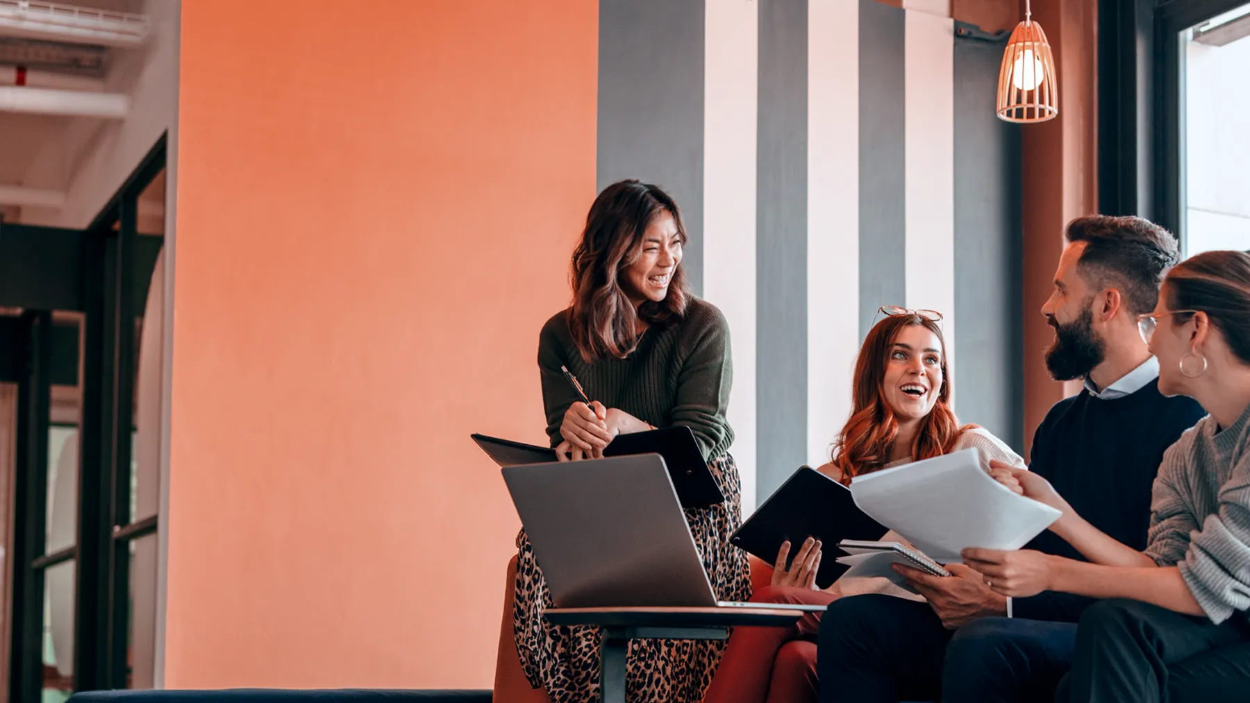 A diverse group of colleagues happily discussing work with laptops, documents, and notebooks in a modern office setting.
