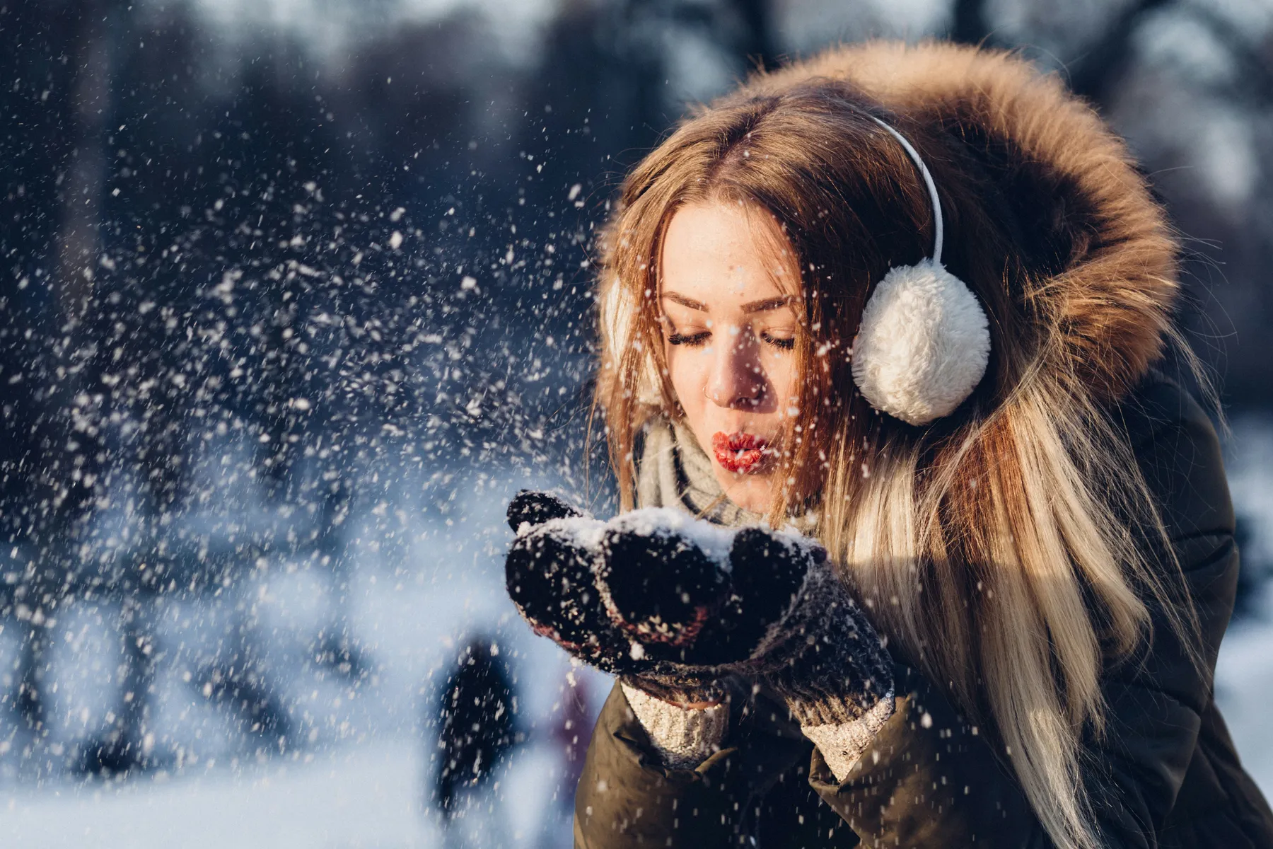A young woman wearing earmuffs and gloves blowing snow from her hands in a snowy outdoor setting.