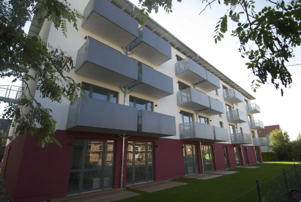 Modern residential building with a red and white facade, featuring large windows and multiple gray balconies arranged in rows. The view is from the garden side, with green lawn and leafy tree branches framing the image.