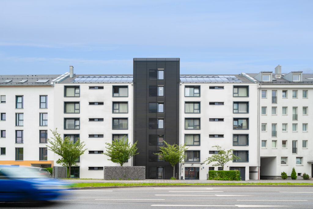 Modern apartment building with a clean white facade and dark central stairwell section, located on a busy street with a blurred blue car passing in the foreground. The building features large windows and small trees planted in front.