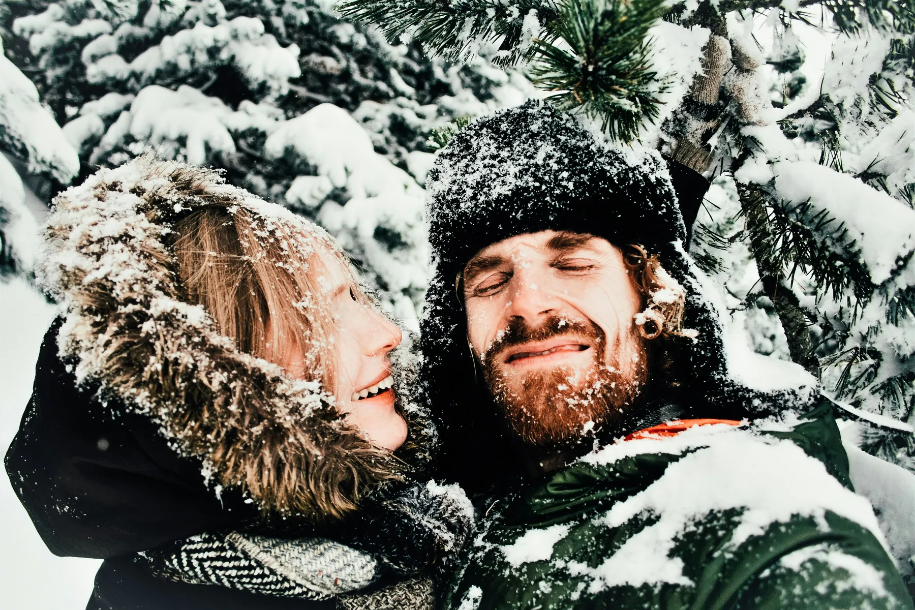 Happy couple taking a selfie in a snowy forest, their faces covered with snow, dressed in warm winter clothes.