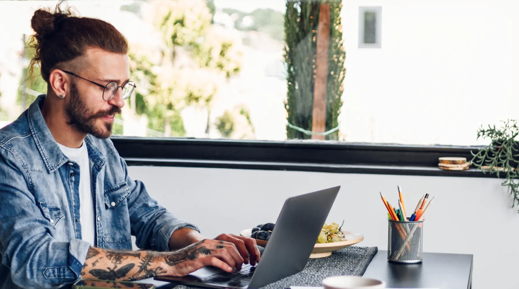 A young man with tattoos and glasses working on a laptop in a bright, modern workspace.