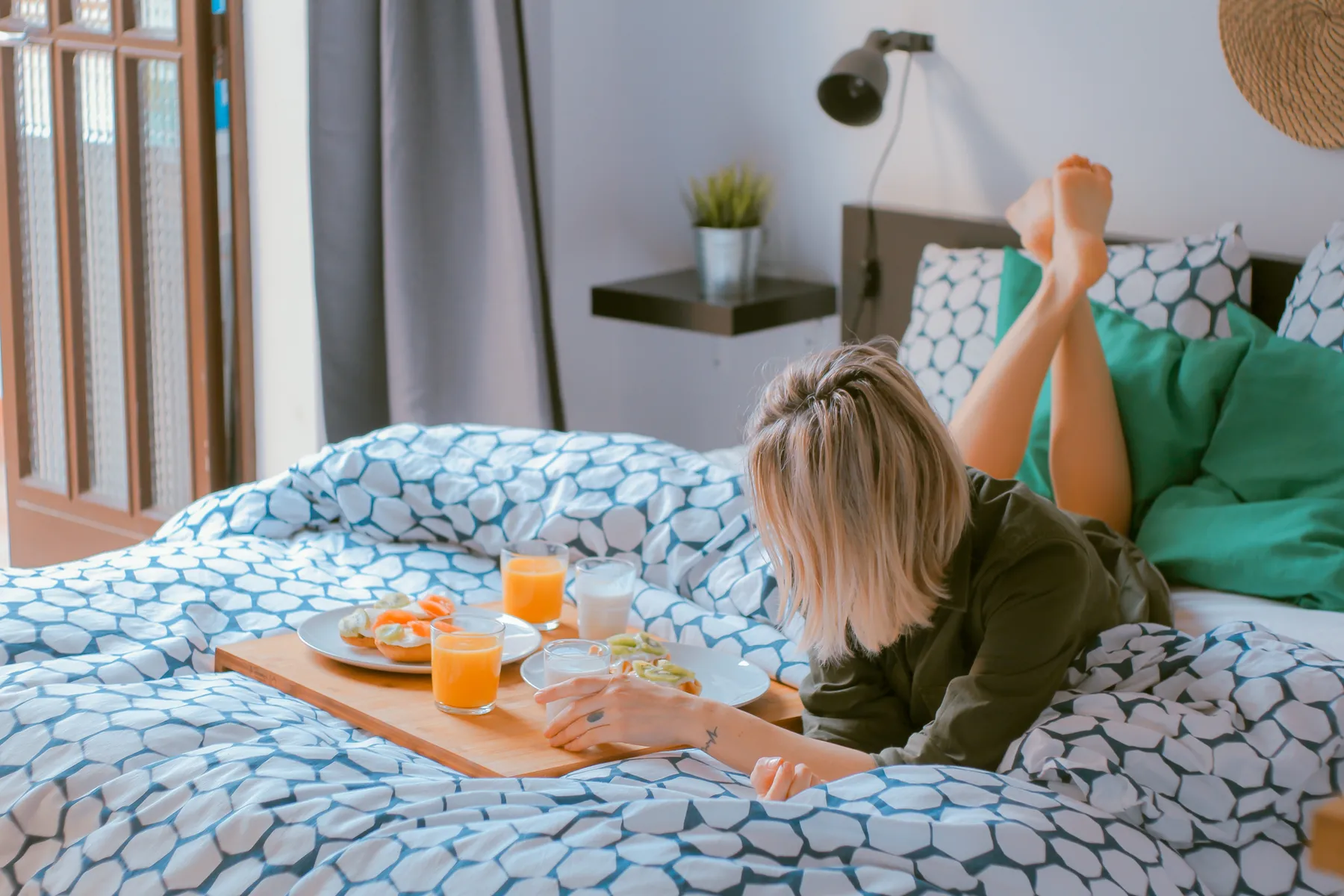 Eine junge Frau liegt entspannt auf einem gemütlichen, gemusterten Bett mit hochgelegten Beinen und genießt ein Frühstückstablett mit Saft, Milch, Obst und Toast. Sanftes Licht und frische Farben vermitteln die Ruhe eines entspannten Morgens zu Hause.