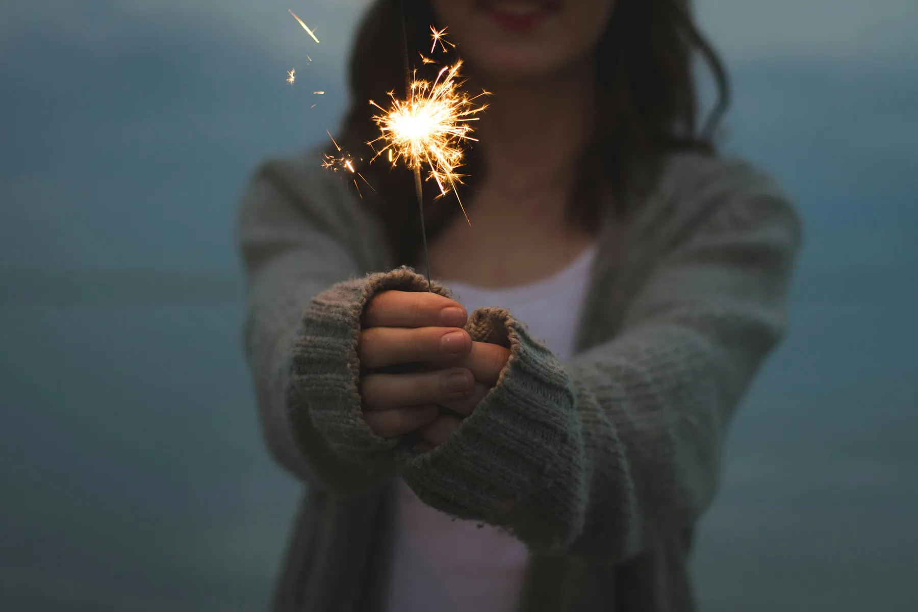 A young woman holds a lit sparkler out in front of her, with her face softly smiling in the background. The sparkler glows brightly against a dim blue backdrop, evoking celebration, warmth, and a quiet, magical moment.