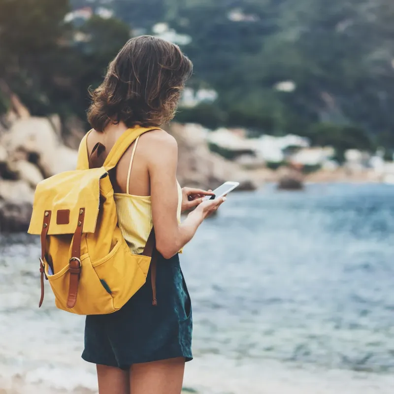 A young woman with a yellow backpack is standing near a rocky coastline, looking at her smartphone. She is dressed in a light top and shorts, with a blurred view of mountains and coastal buildings in the background.