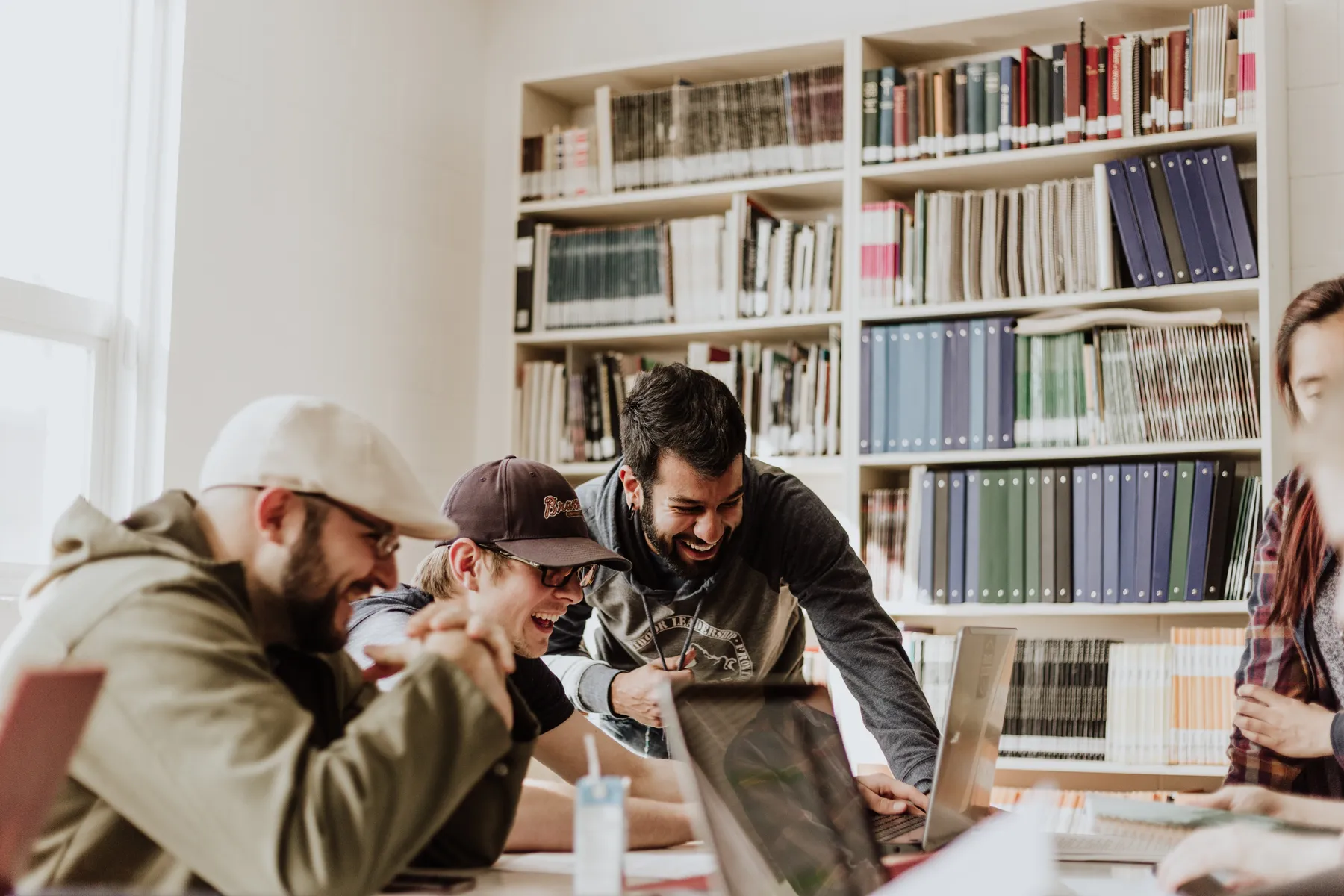 Eine Gruppe junger Erwachsener lacht und arbeitet gemeinsam an Laptops in einer hellen Bibliothek oder Lernumgebung. Umgeben von Bücherregalen, scheinen sie ein gemeinsames Projekt oder einen Witz zu genießen – Lernen trifft auf Spaß.