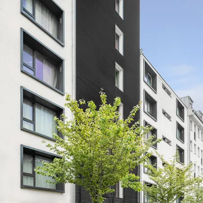 Modern apartment building with a combination of white and dark grey facade panels. The building features large windows and is lined with green trees in front, under a clear blue sky.