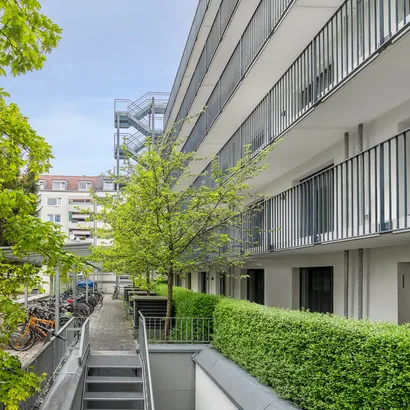 Modern apartment building with balconies and a landscaped courtyard. The courtyard features green hedges, young trees, and a row of parked bicycles along a walkway.