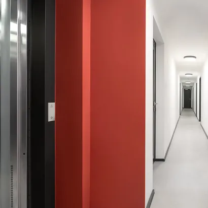 Modern interior hallway of an apartment building, with white walls, black trim, and a striking red accent wall near an elevator.