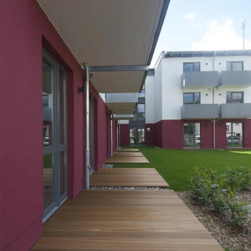 Ground-level view of a modern apartment complex with burgundy and white walls. Wooden terraces extend from each unit, bordered by a neatly maintained green lawn and young plants.