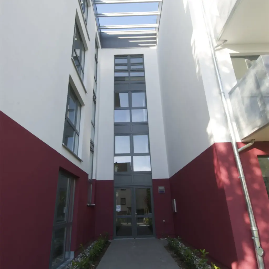 View into the courtyard of a modern residential building with clean architectural lines. The facade is painted white and deep red, featuring large windows and a glass canopy above that allows natural light into the entrance area.