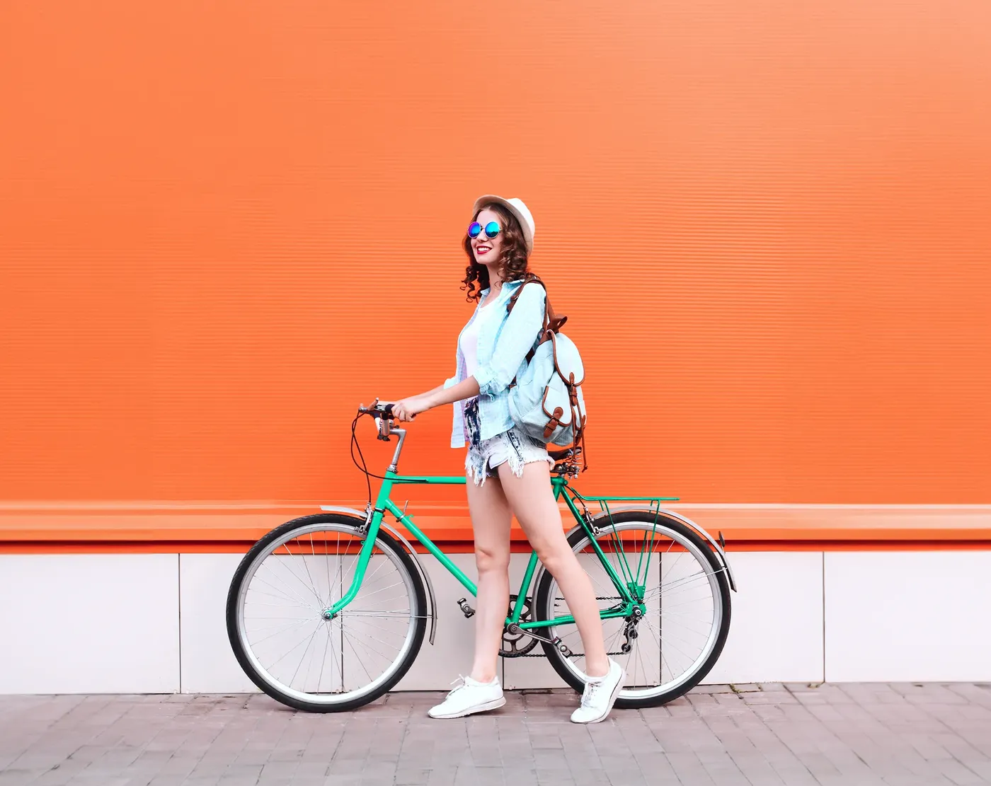 Young woman wearing sunglasses, a hat, and a backpack standing with a green bicycle in front of a bright orange wall.